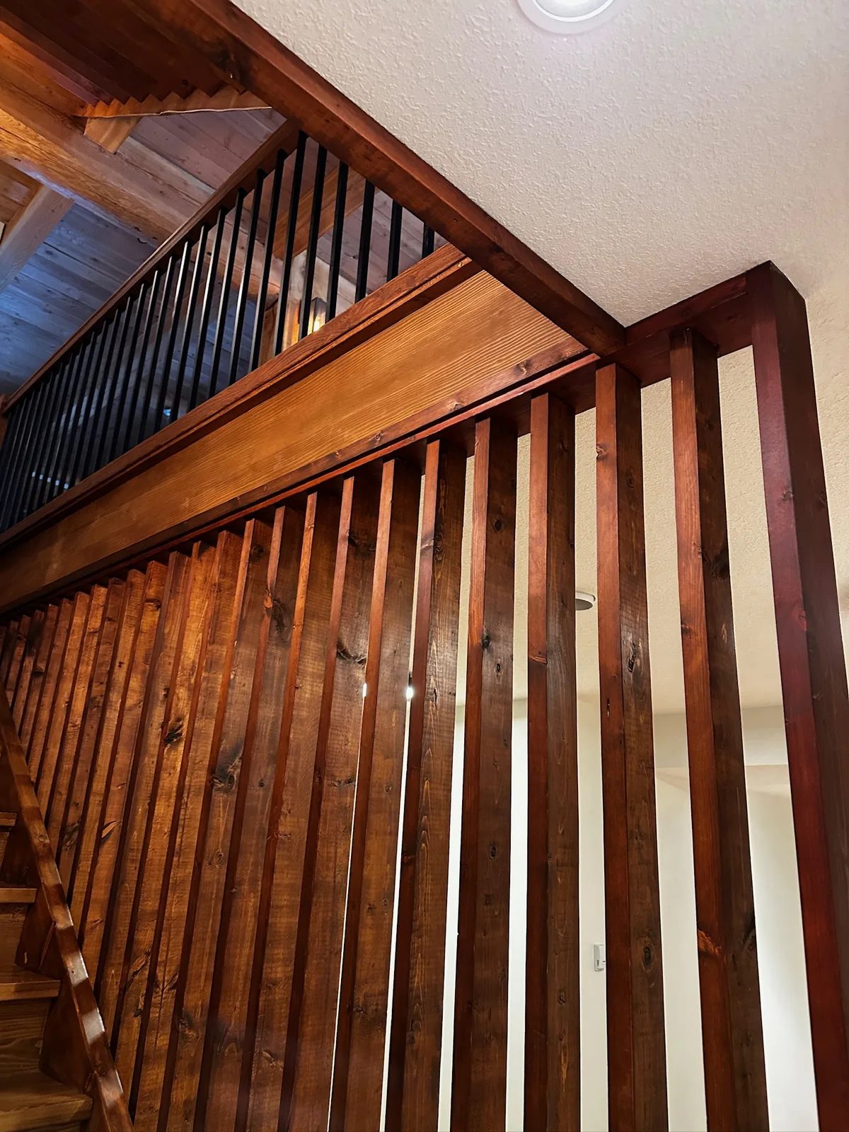 Stained wood slat railing under a sloped pine ceiling beside a staircase